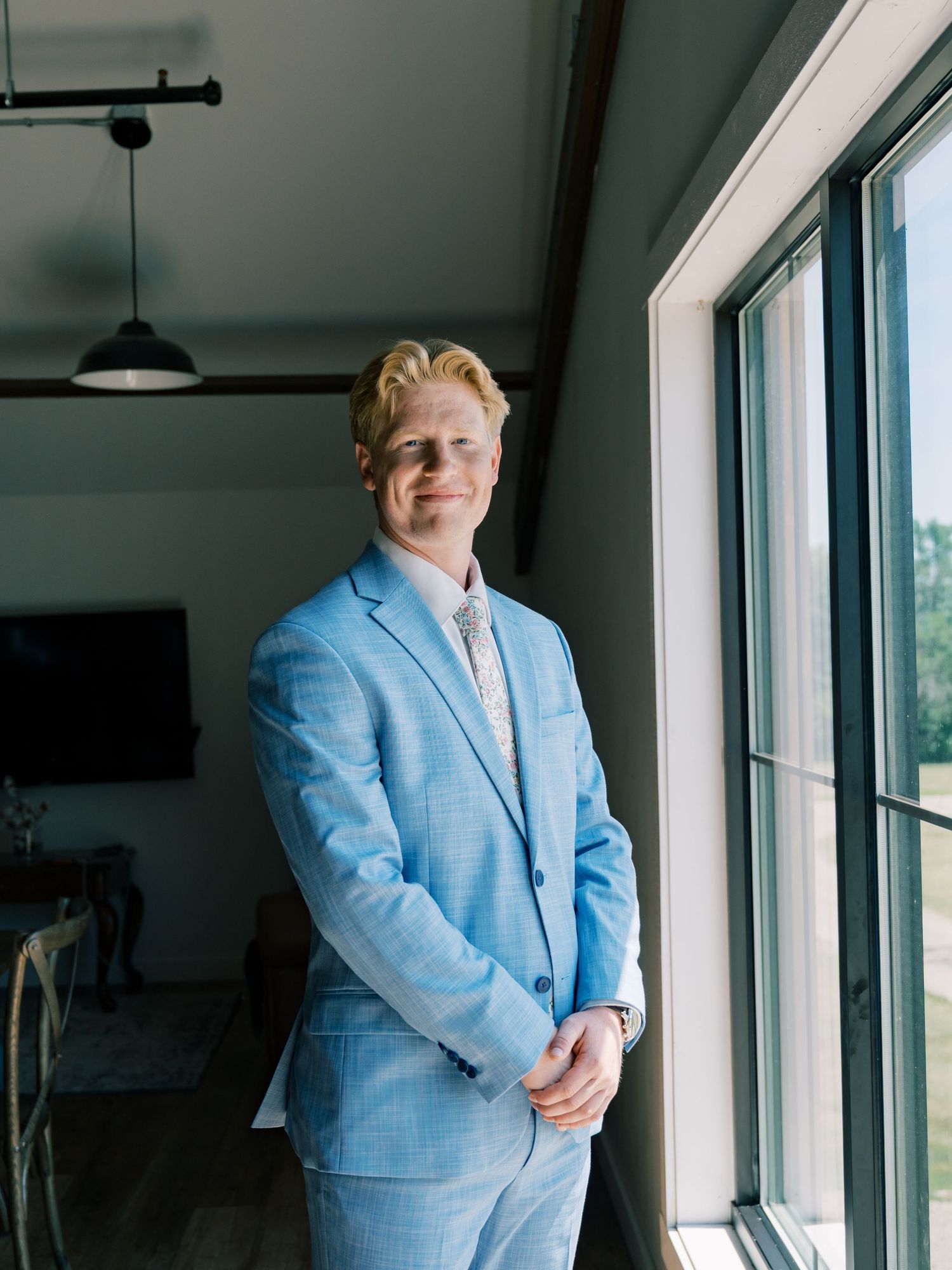 A young man in a light blue suit stands by a large window in a modern room, smiling confidently.