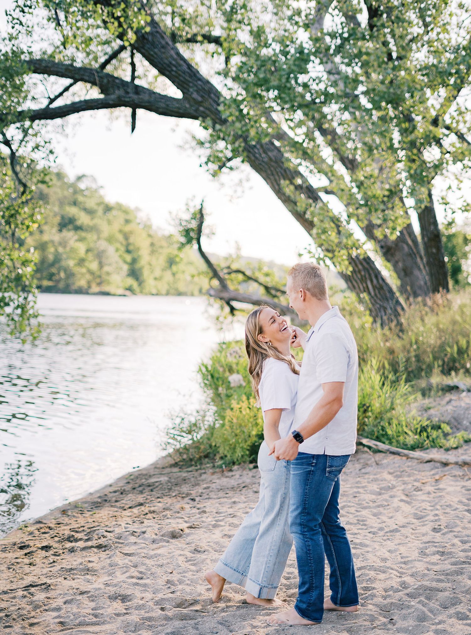 A couple embraces on a sandy beach near the water under large overhanging trees on a sunny summer day.