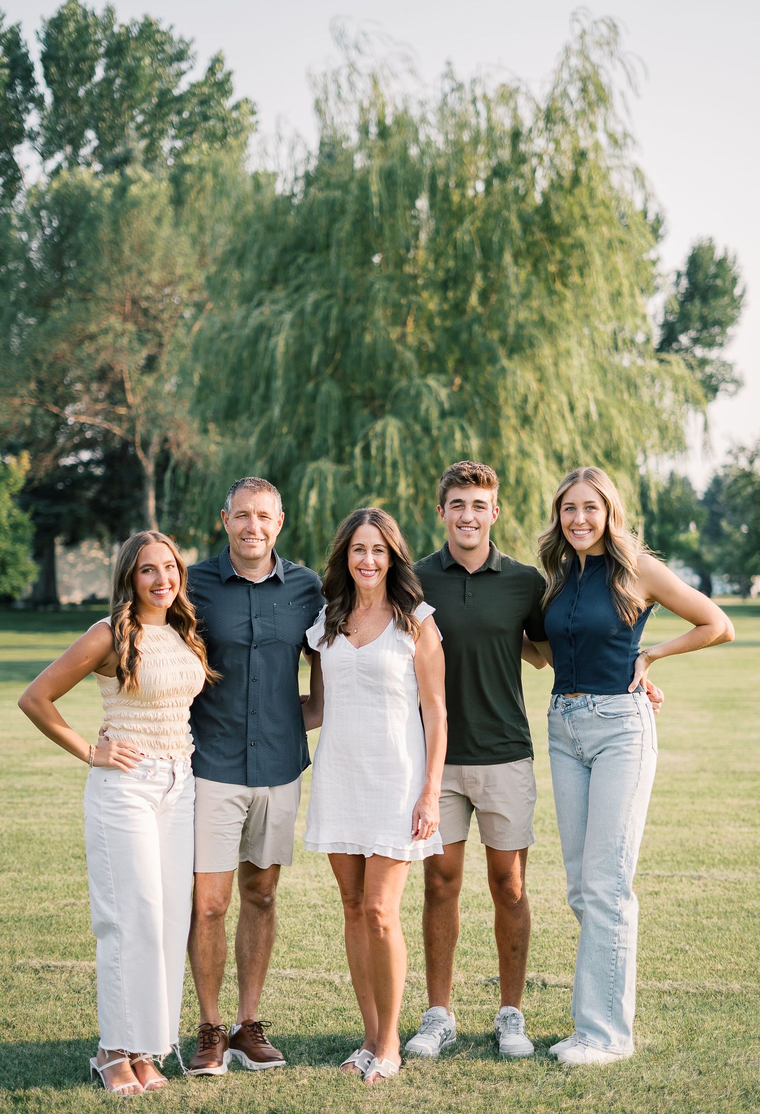 A group of five people standing together outdoors on grass with a willow tree in the background on a sunny day.