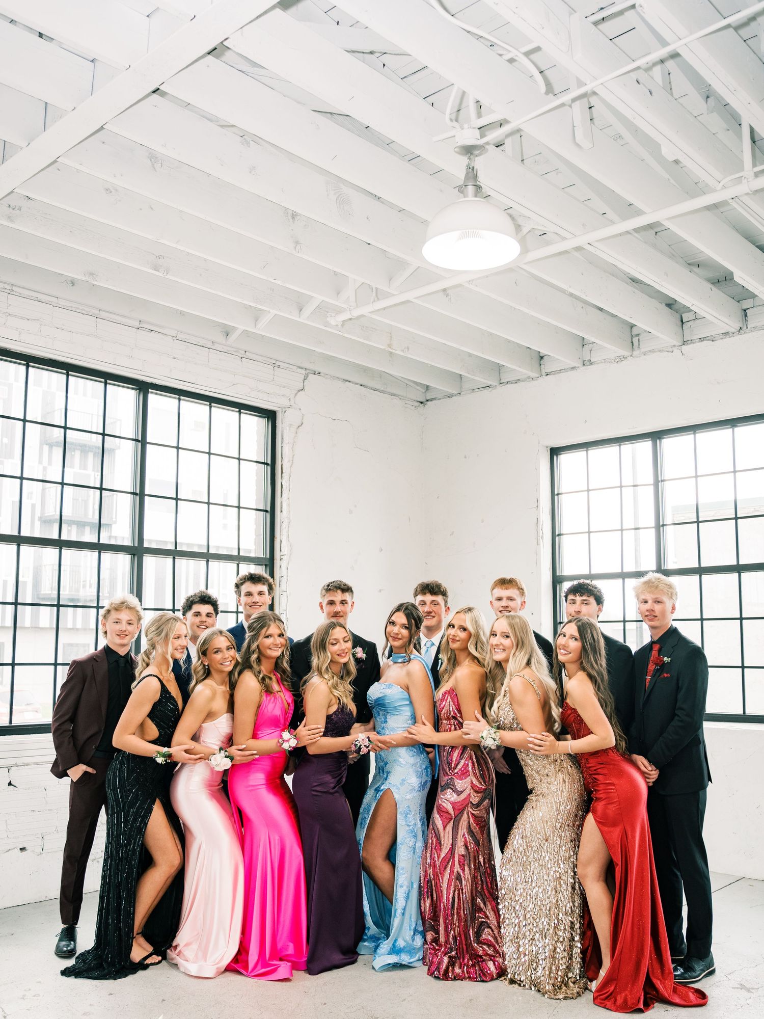 Group of teens in formal prom attire, girls in colorful gowns and boys in suits, posing together in a bright white venue.
