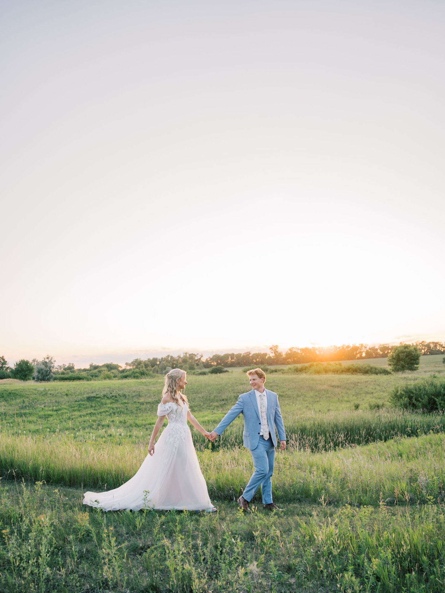 A bride and groom hold hands walking through a lush green field at golden hour sunset on their wedding day.