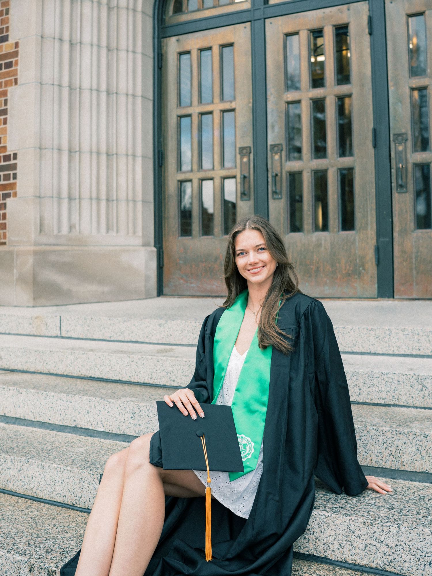 Young woman in black graduation gown and cap sitting on steps outside a university building, smiling.