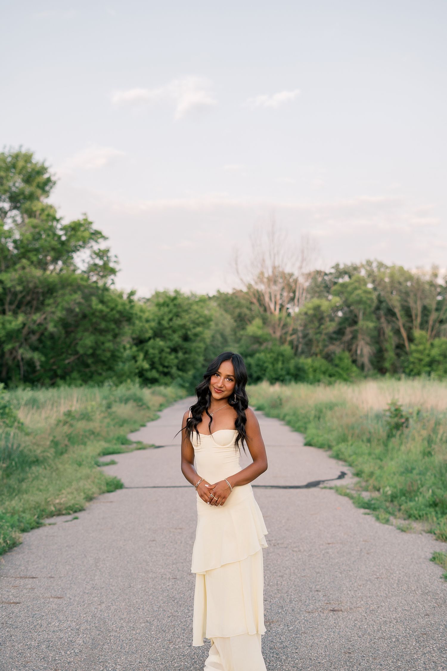 A person in a flowing white dress poses gracefully on a gravel path surrounded by lush greenery on a sunny day.