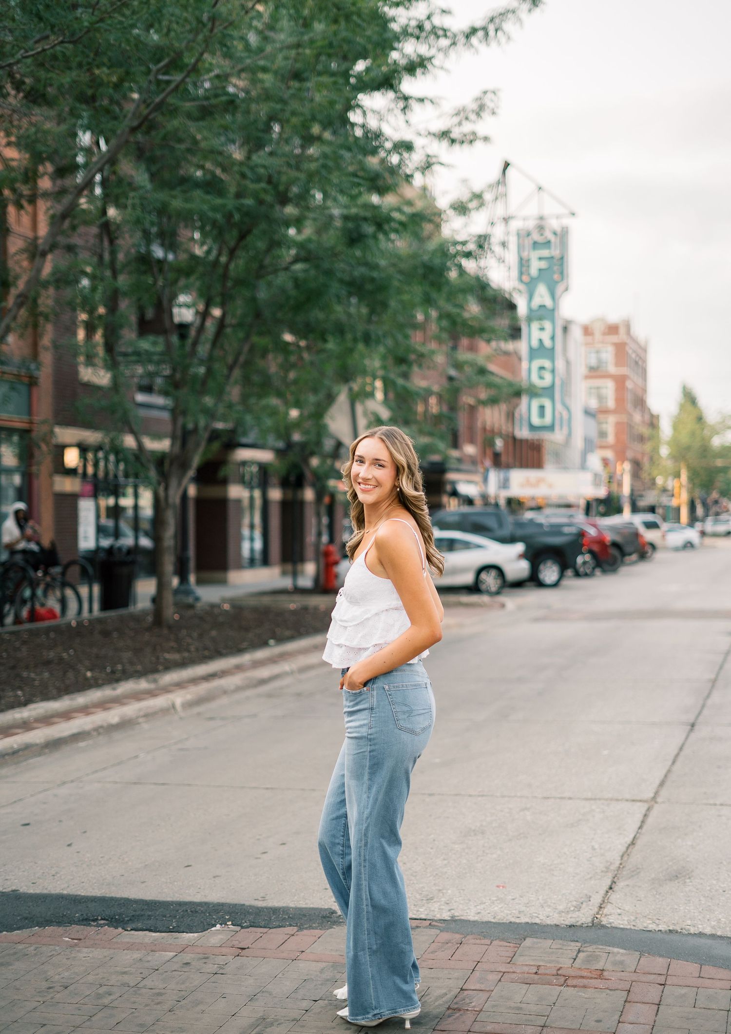 A person stands on a city street wearing light wash flare jeans and a white top, with brick buildings and trees in view.