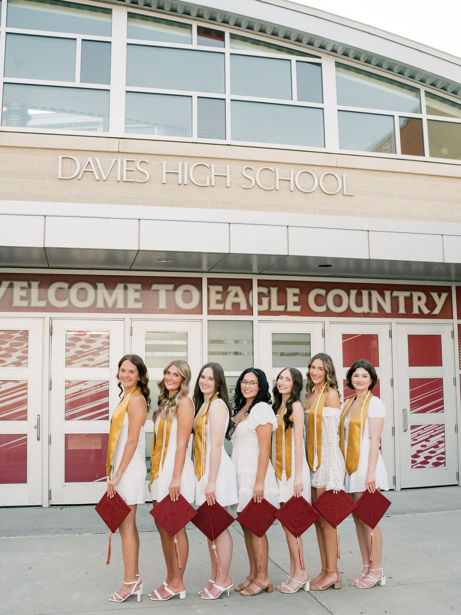 Eight female graduates in white dresses holding burgundy caps pose outside Davies High School Welcome to Eagle Country sign.
