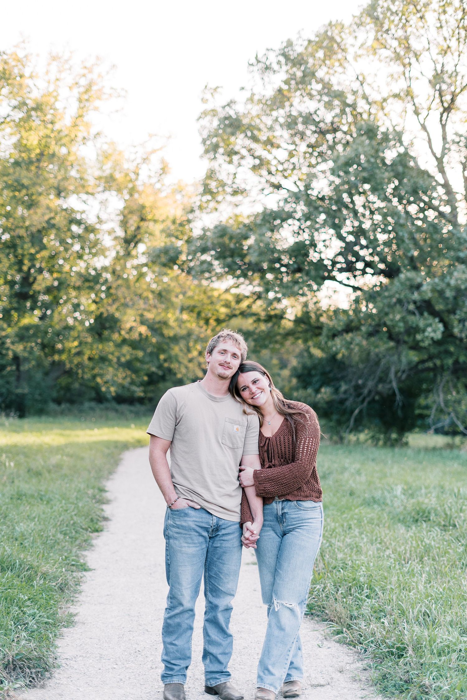 A couple stands together on a sunlit nature trail surrounded by green trees and grass on a warm afternoon.