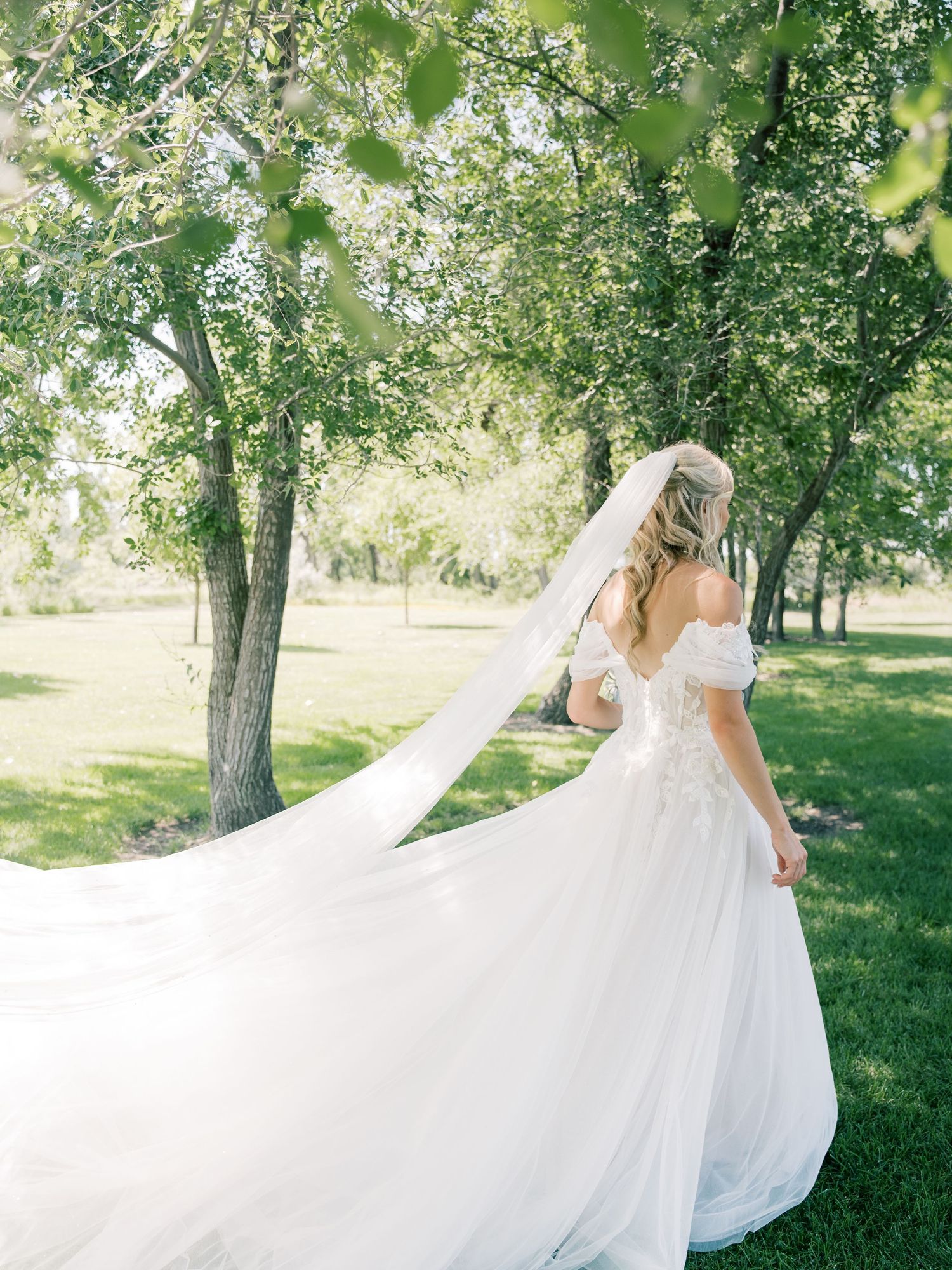 Bride in flowing white off-shoulder gown with cathedral veil billowing in breeze among lush green trees.