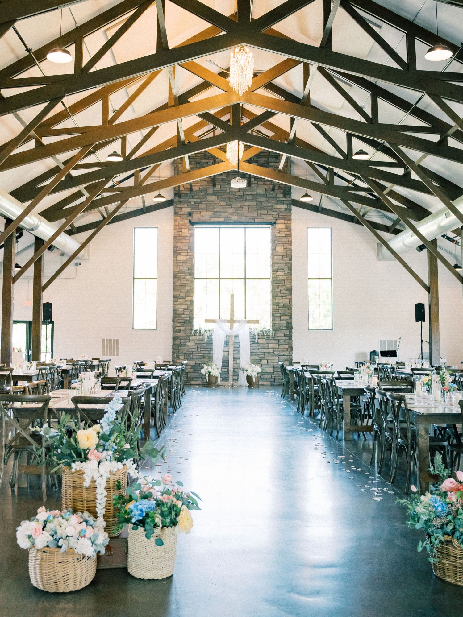 Rustic wedding venue with vaulted wood beam ceiling, floral basket aisle decor, and elegant reception tables in a barn-style hall.