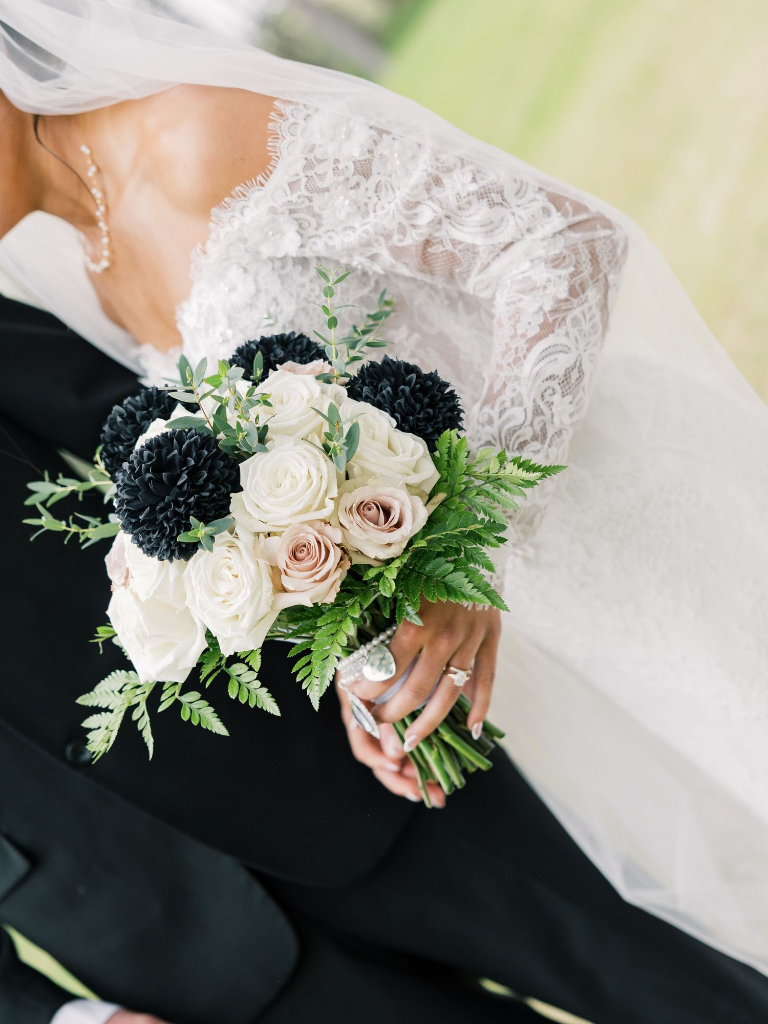 Bride holding a wedding bouquet with cream roses, black dahlias, and greenery, wearing a lace gown and diamond ring.