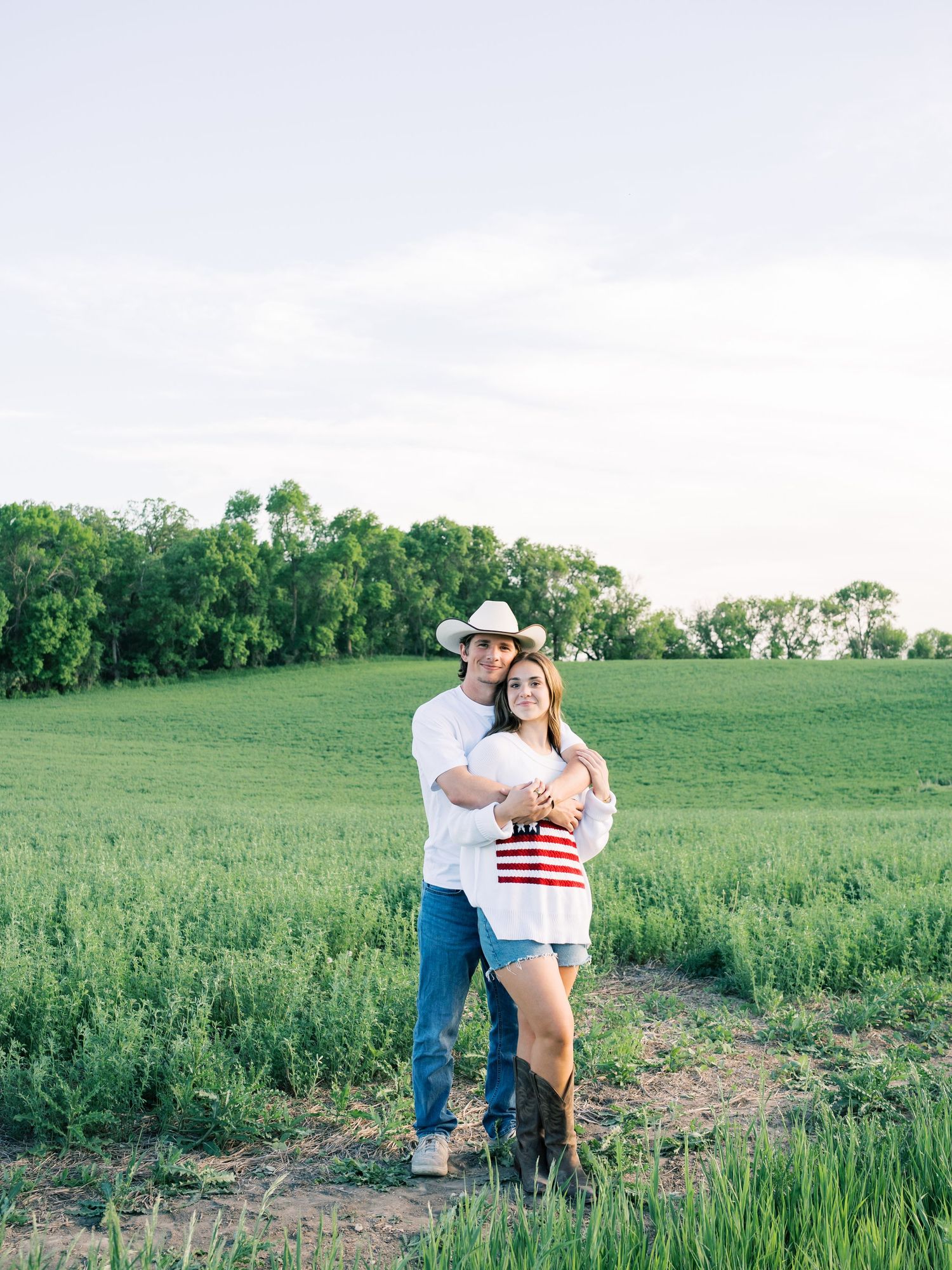 Couple embracing in a green field, wearing cowboy hats and denim, during a romantic country engagement photo session.