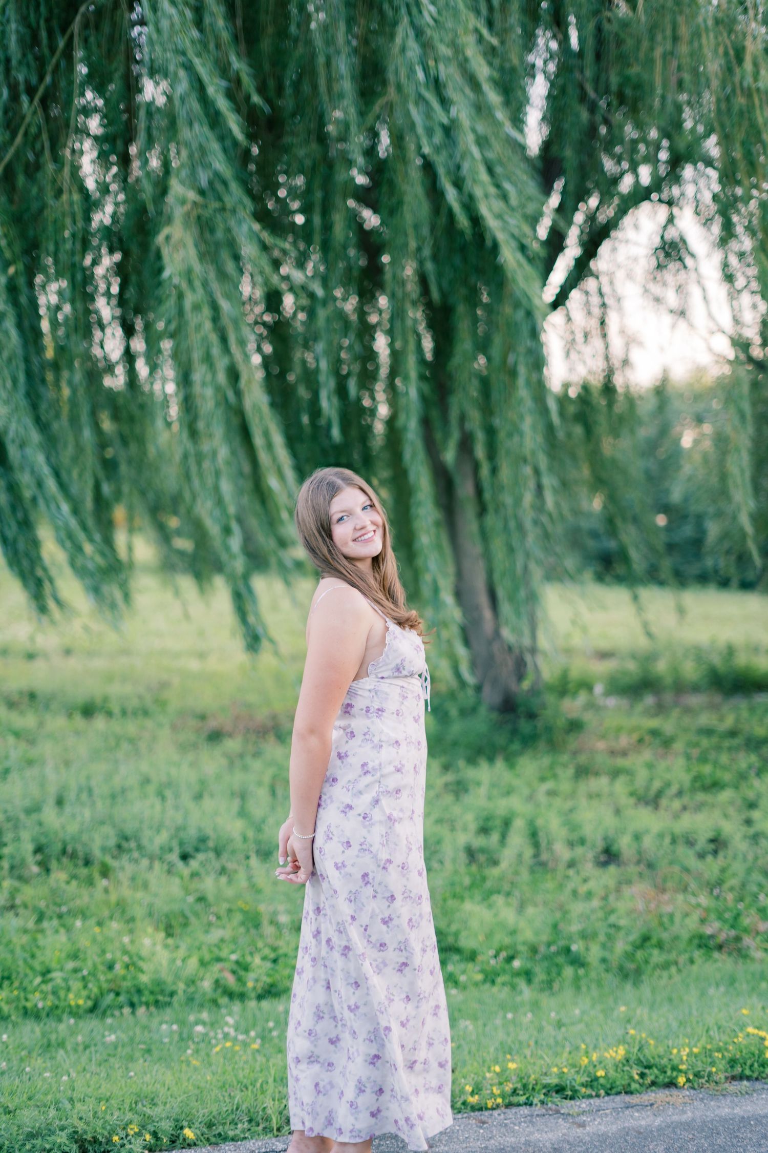 Woman in a floral sundress poses gracefully under drooping willow tree branches in a serene outdoor setting.