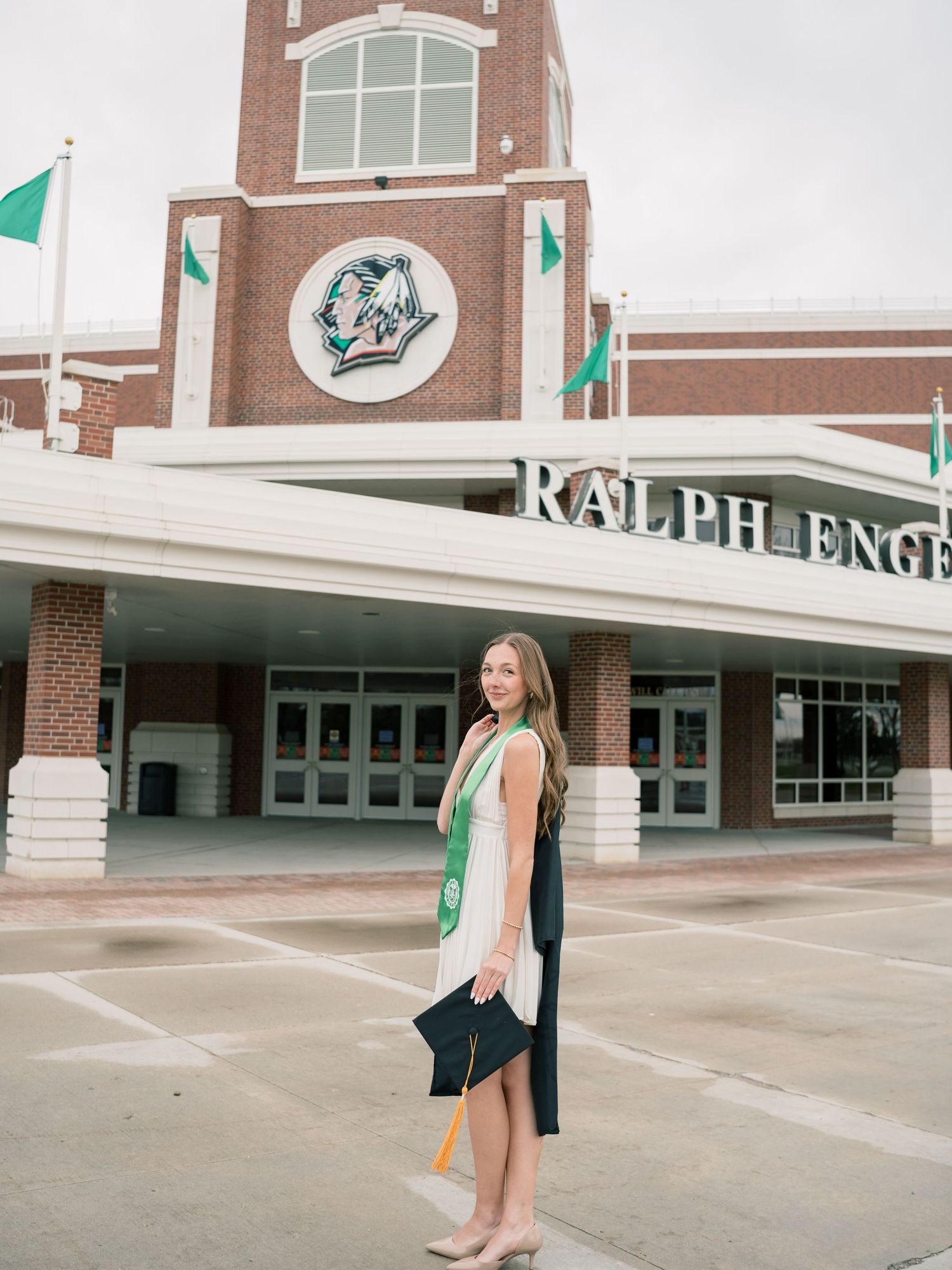 A young woman in graduation attire stands proudly outside the Ralph Engelstad Arena, wearing a green stole.