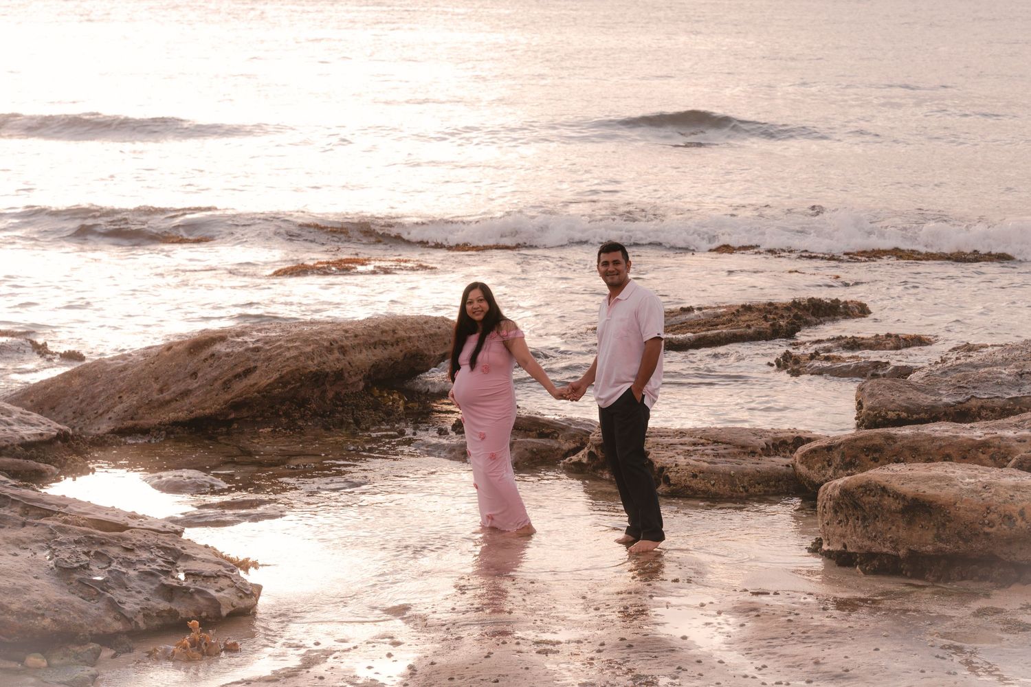 Couple holding hands on rocky beach shore at sunset, woman in pink maternity dress, romantic seaside maternity photo.