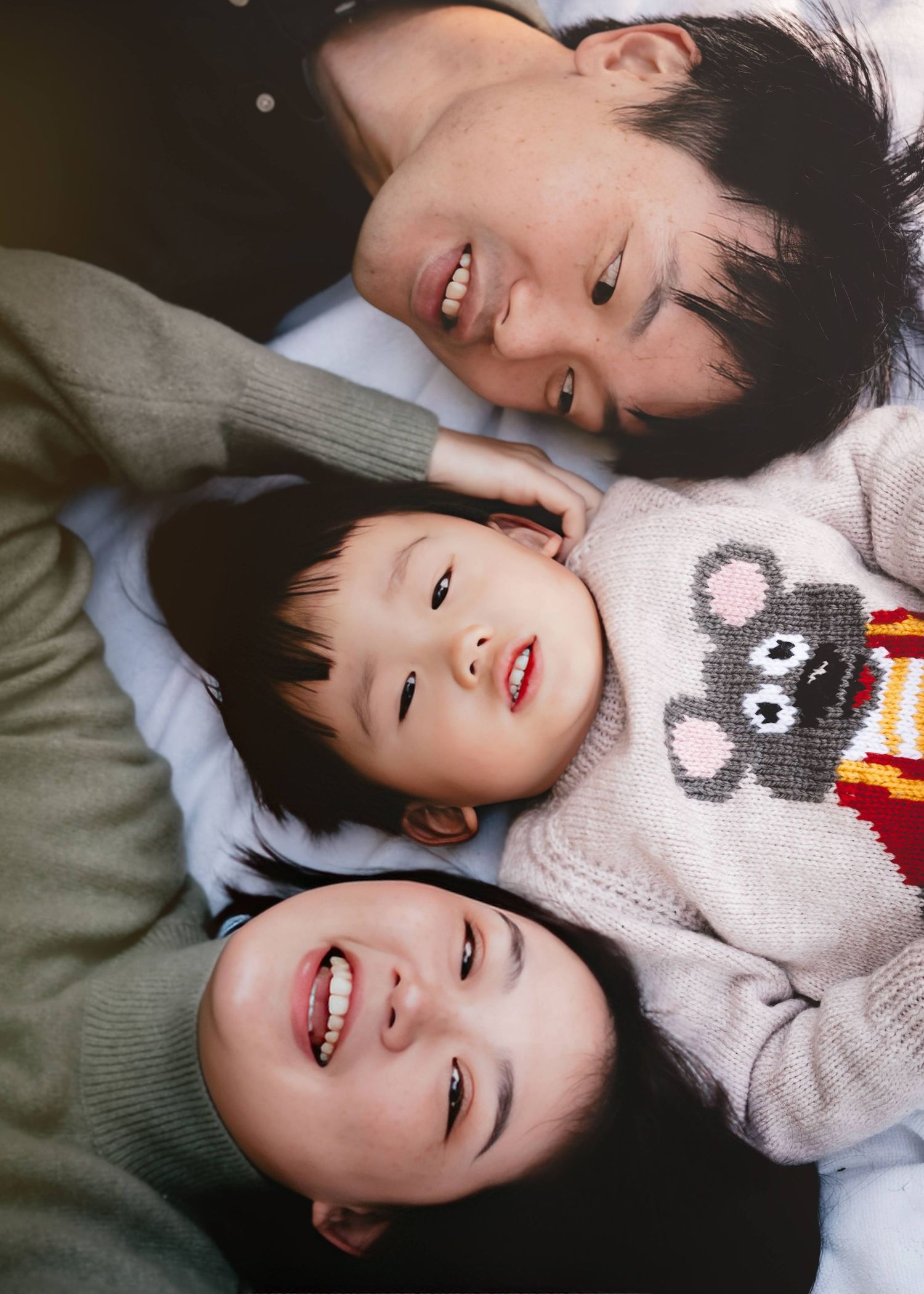 Three smiling children lie down together wearing sweaters in a candid photo.
