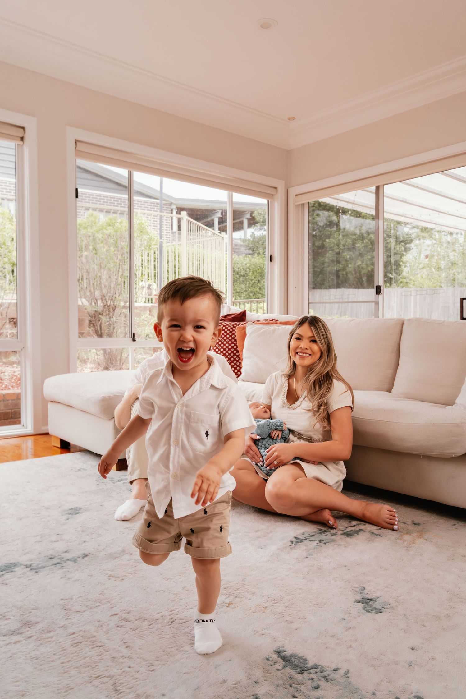 A joyful toddler boy runs laughing in a bright living room while his smiling mother sits on a rug nearby.