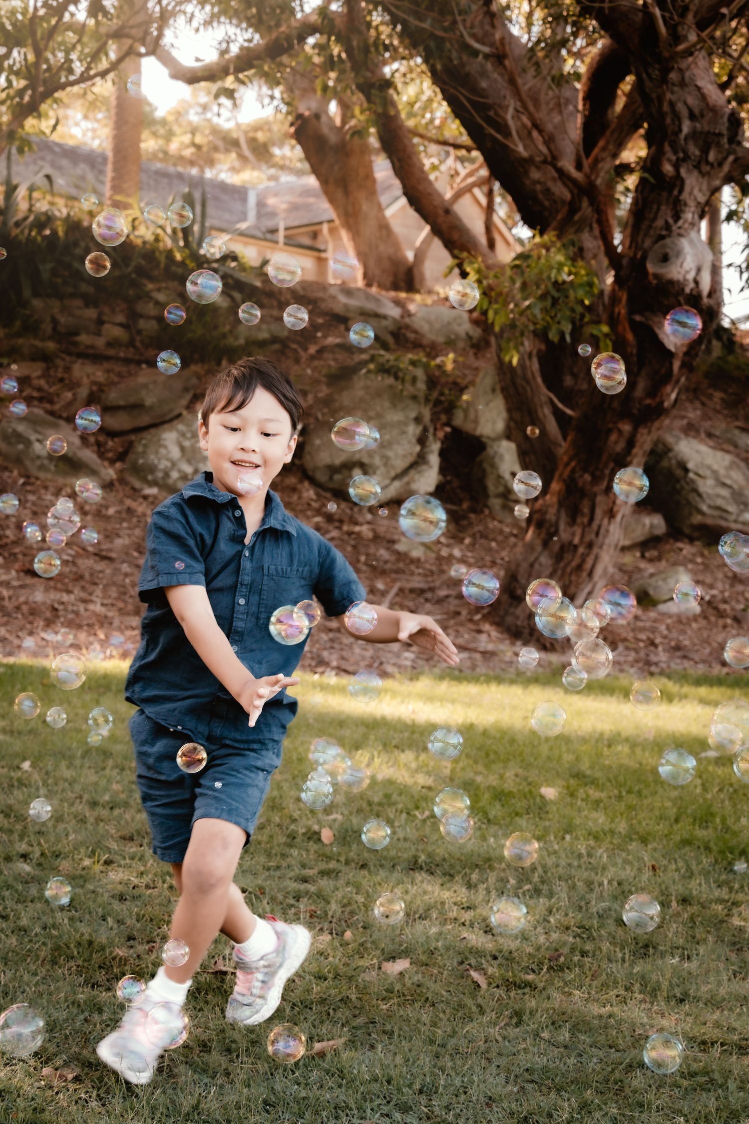 A joyful young boy in denim outfit plays with bubbles outdoors in a sunny park setting.