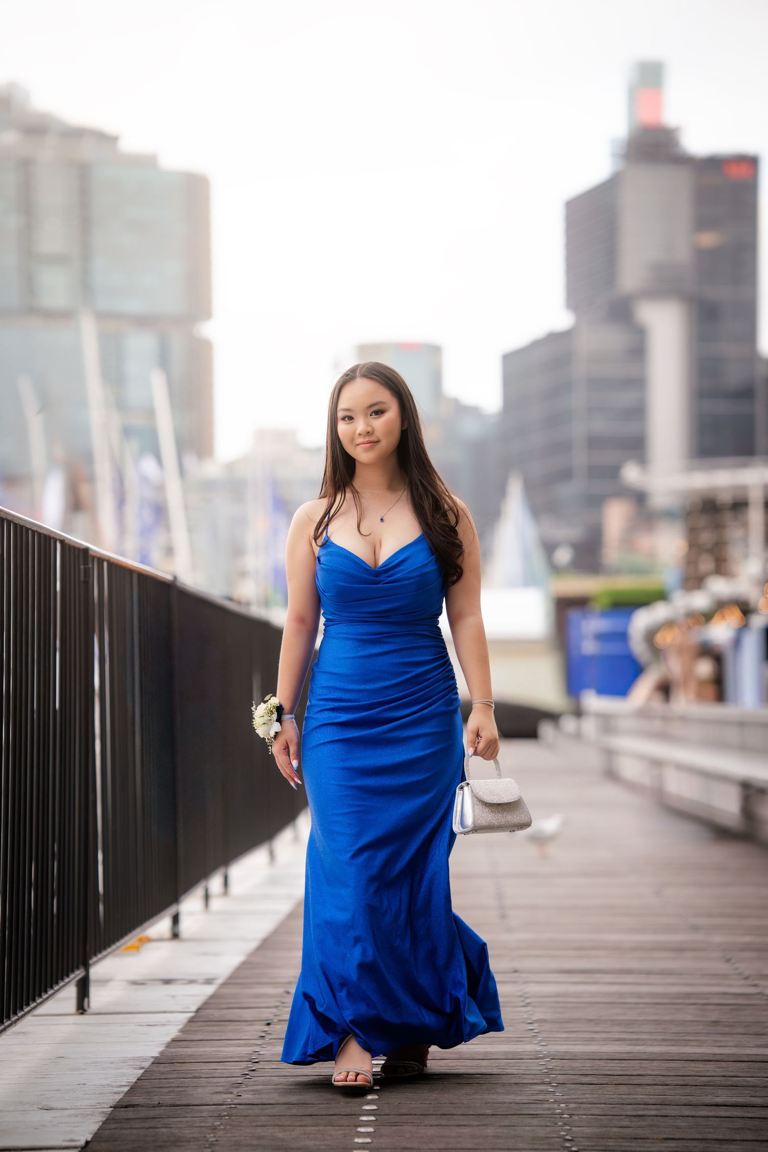 A woman in a vibrant blue dress poses on a wooden boardwalk with city buildings in the background.