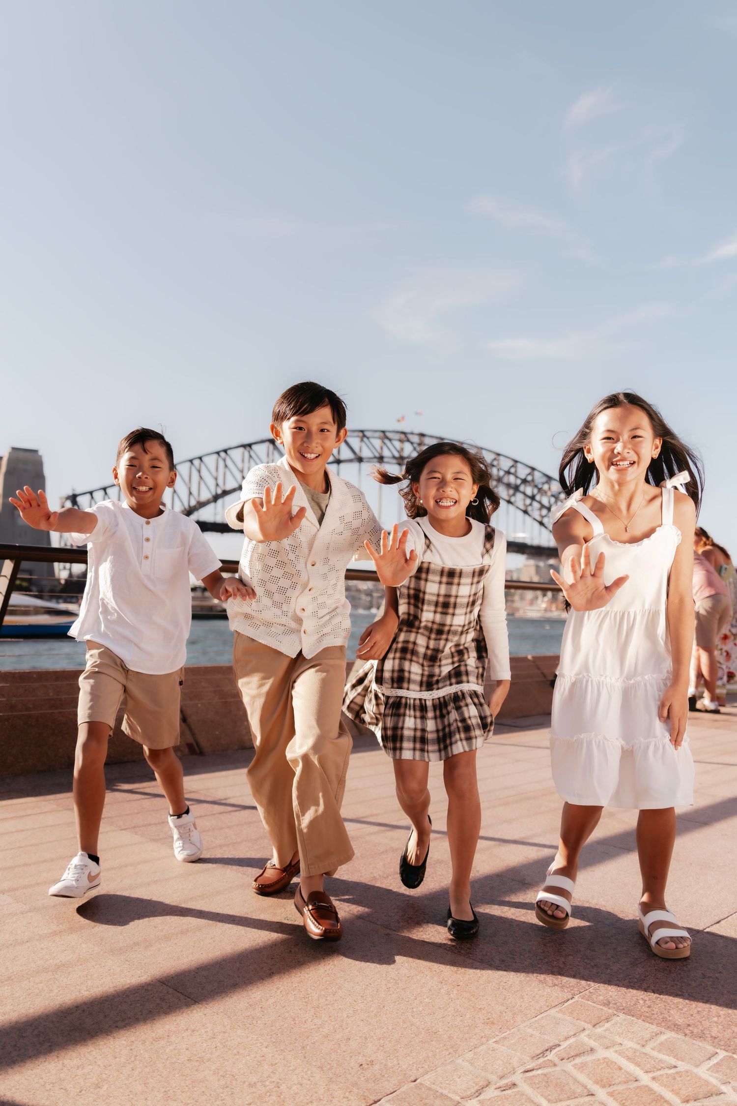 Four children in coordinated white and plaid outfits run and play together on a boardwalk by the water.