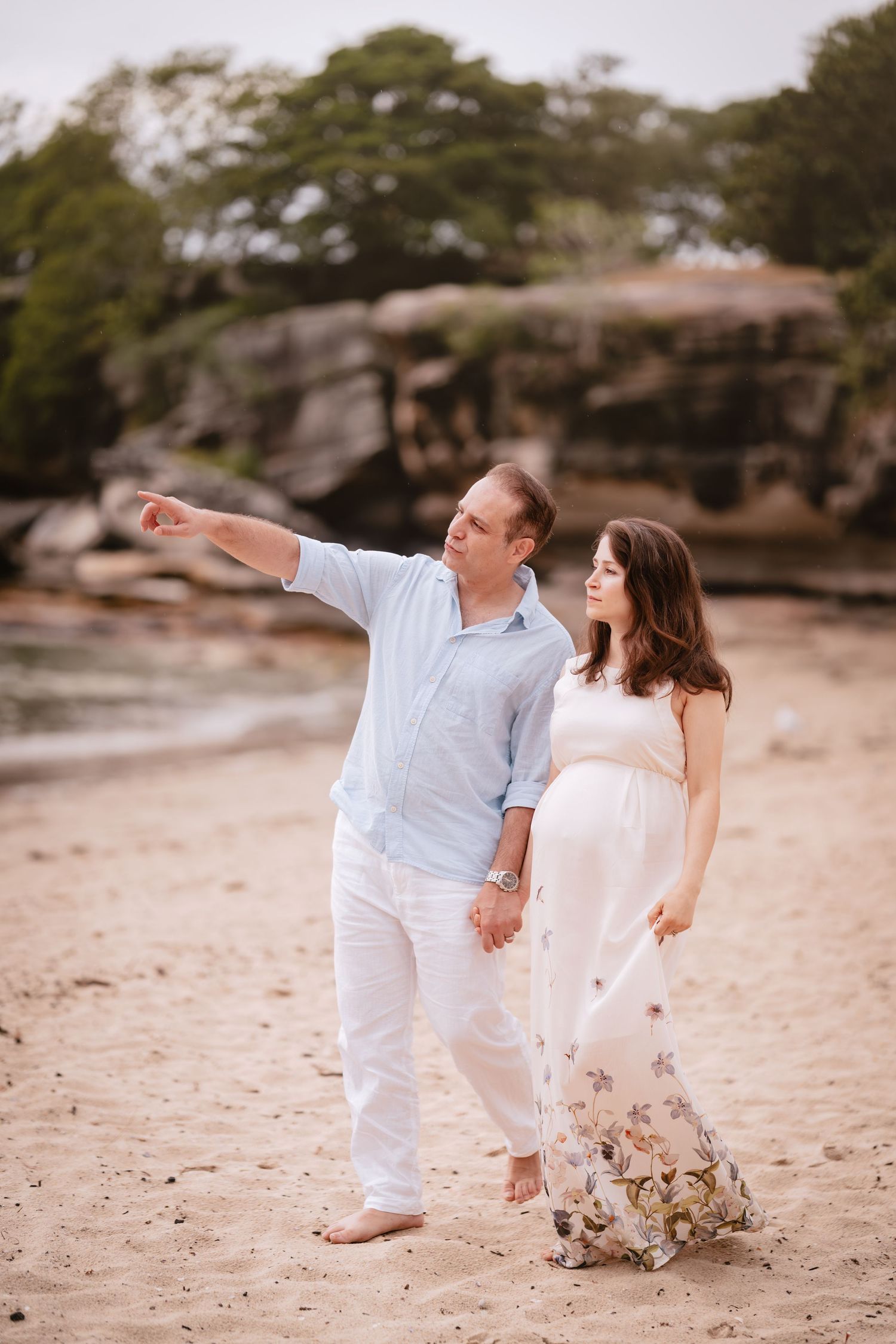 A couple in white outfits walking playfully along a sandy beach with rocks in the background.