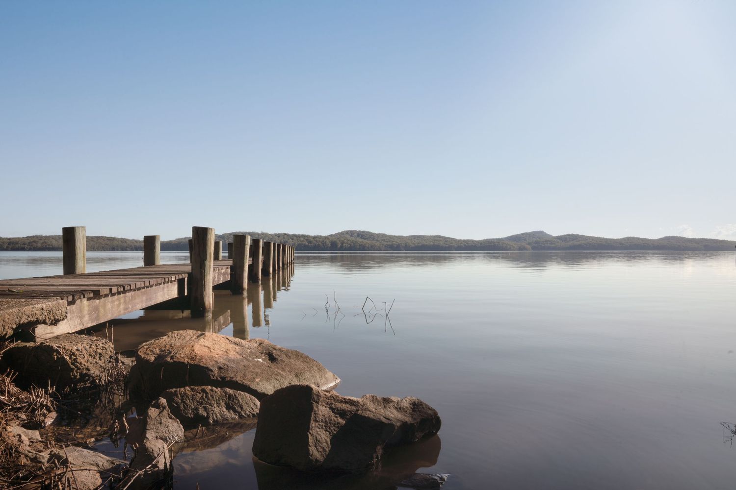 A long wooden pier extends across a tranquil lake at sunrise, with mountains visible in the misty background.