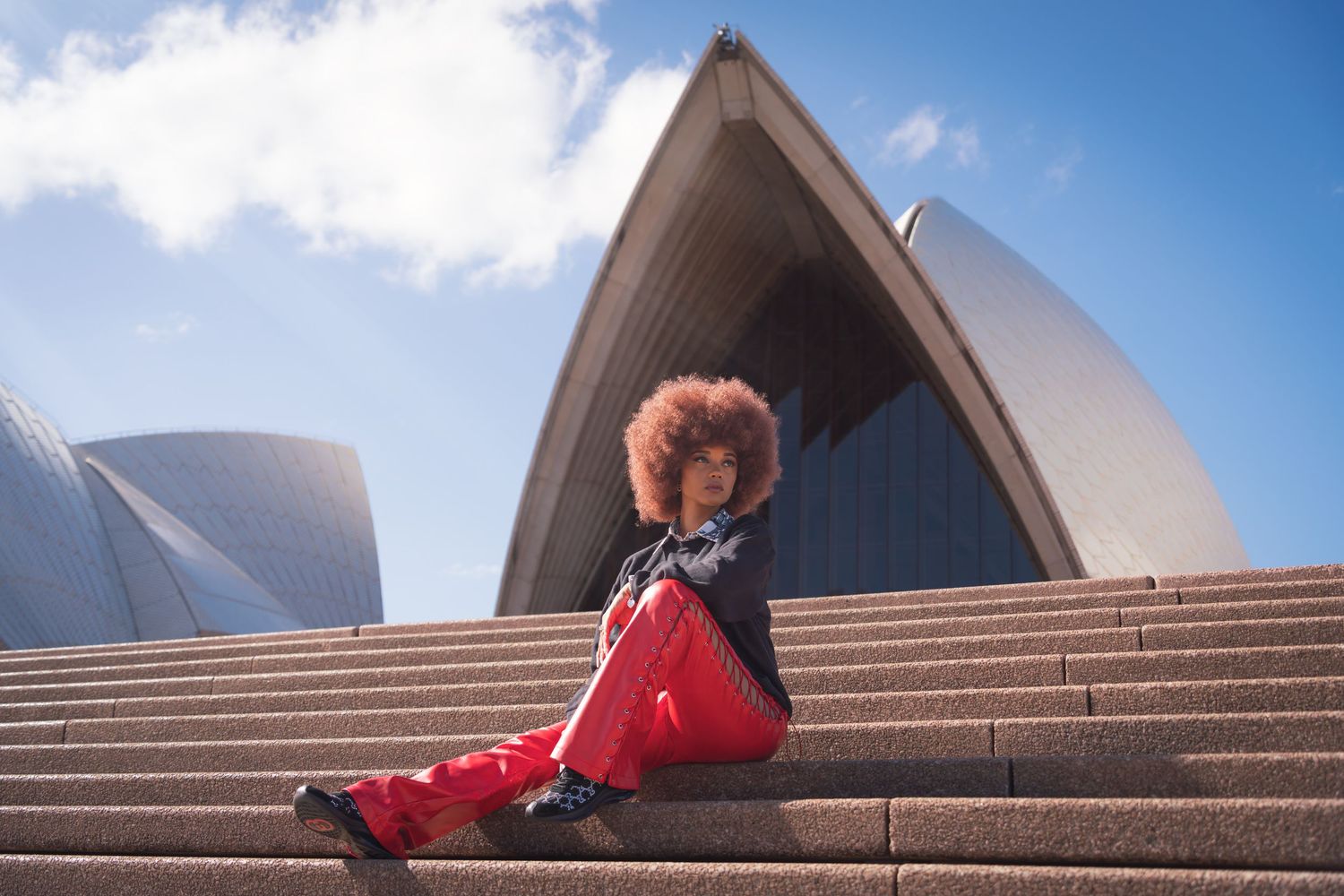 A person in a red outfit sits on the iconic steps of the Sydney Opera House on a sunny day.
