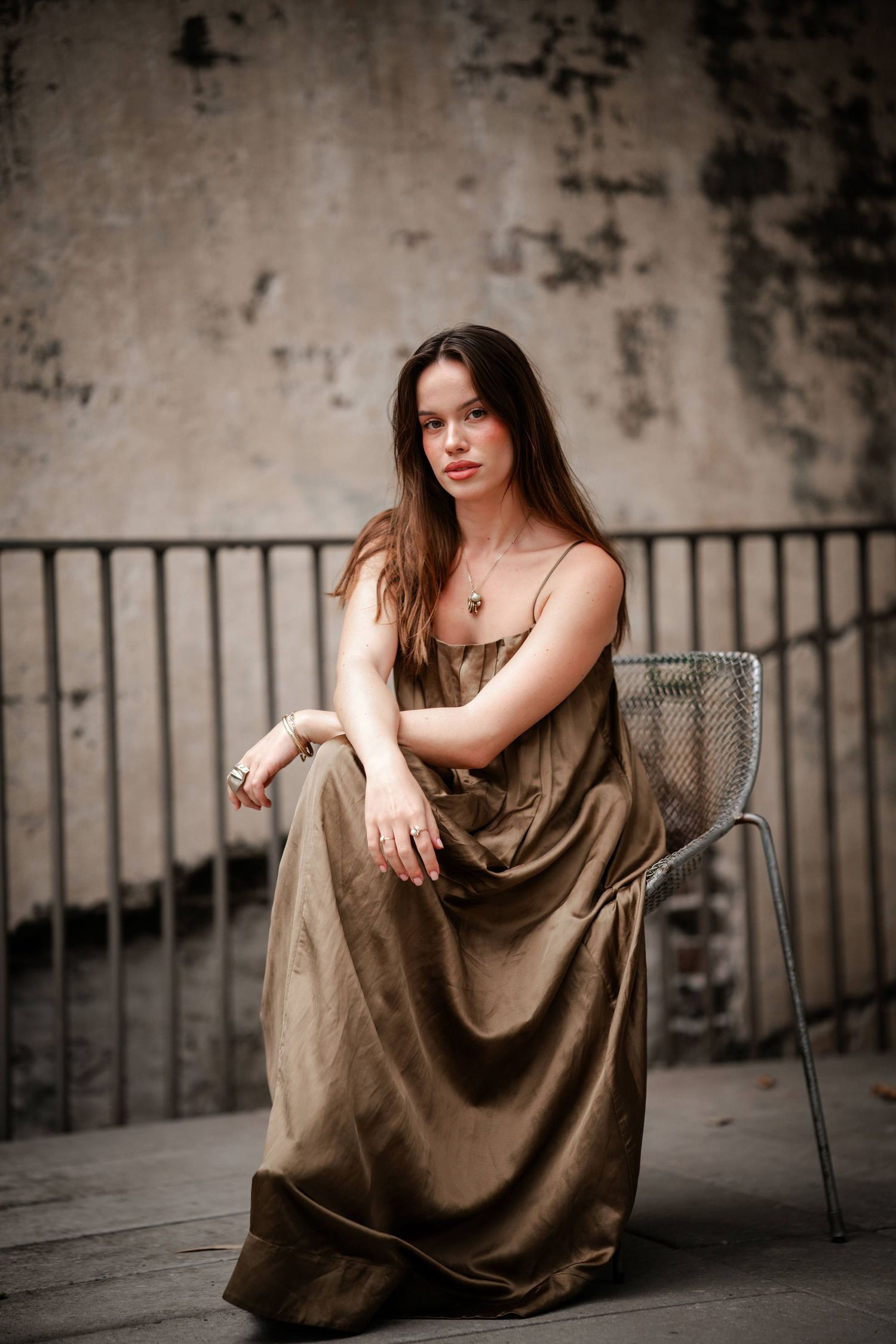 A figure in a flowing brown dress poses gracefully on a chair against a textured concrete wall.