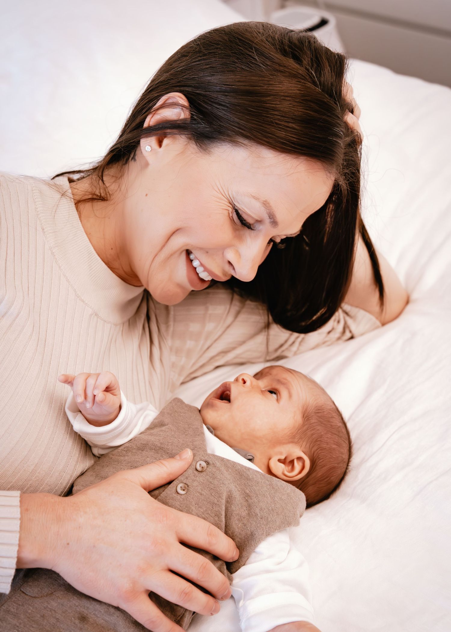 A mother and baby share tender moments together while lying on white bedding.