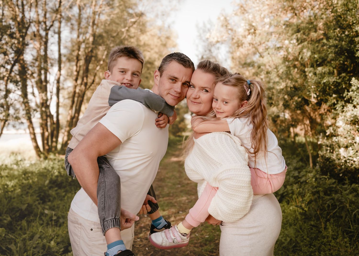 A family wearing cream and pink outfits embraces outdoors during a sunset photoshoot in a wooded setting.