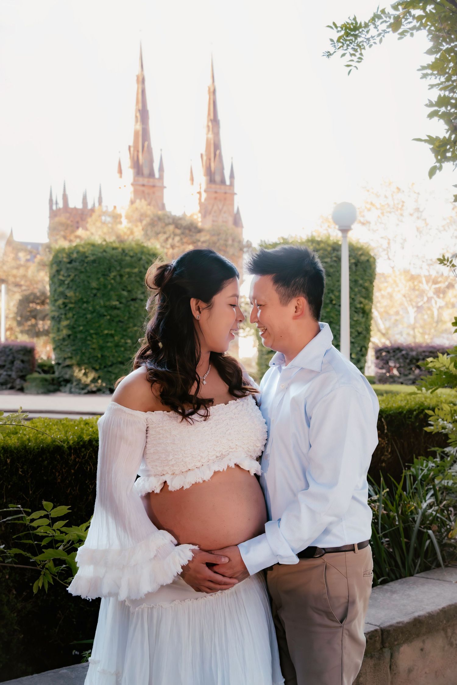 Pregnant couple sharing a tender moment outdoors, with Gothic church spires and lush hedges in the background.