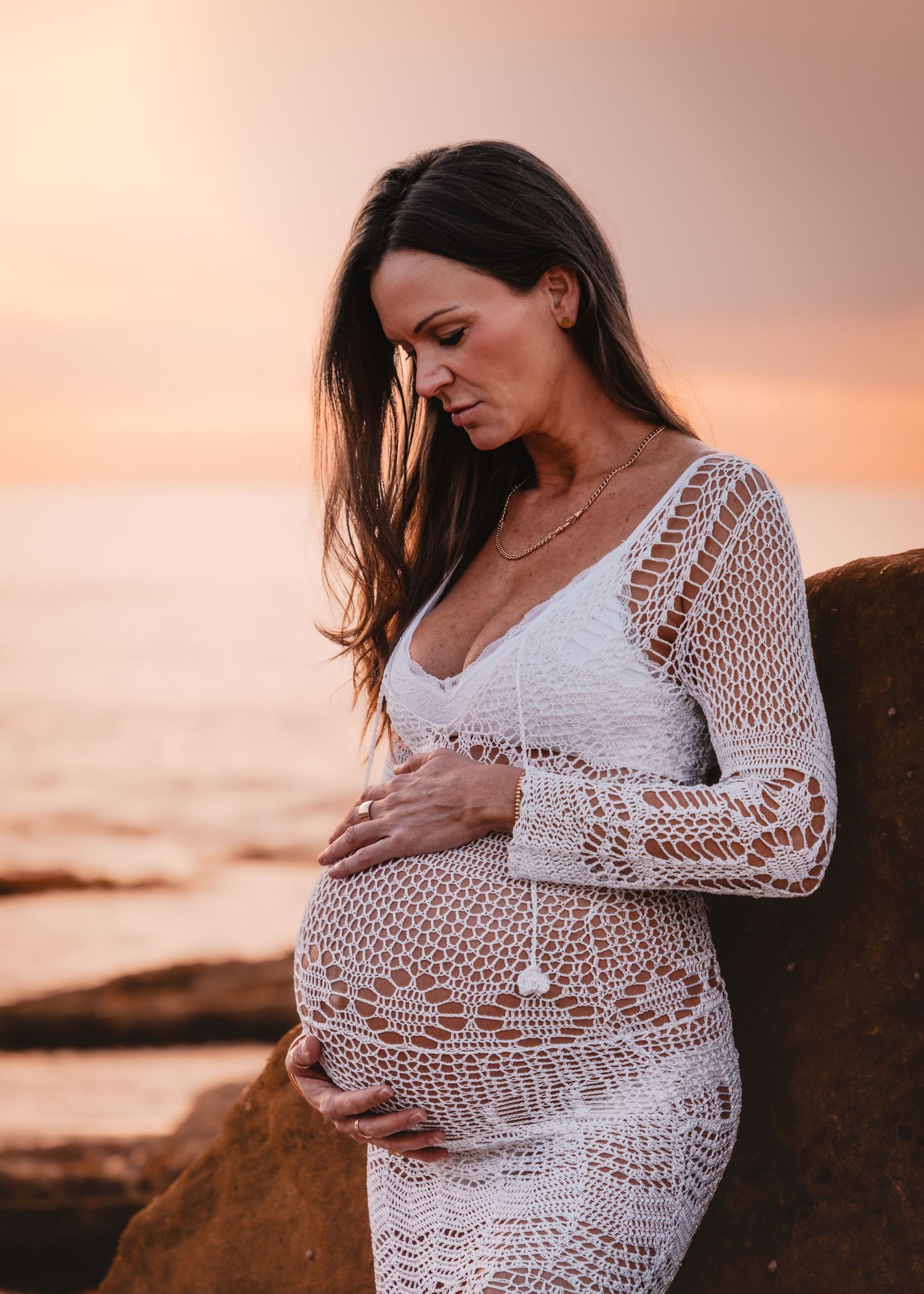 A person in a white lace dress cradles their pregnant belly against a warm sunset backdrop by the ocean.