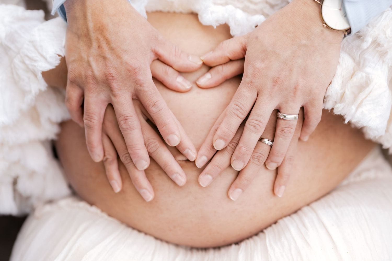 Two pairs of hands forming a heart shape on a pregnant woman's bare belly, symbolizing love and expectancy.