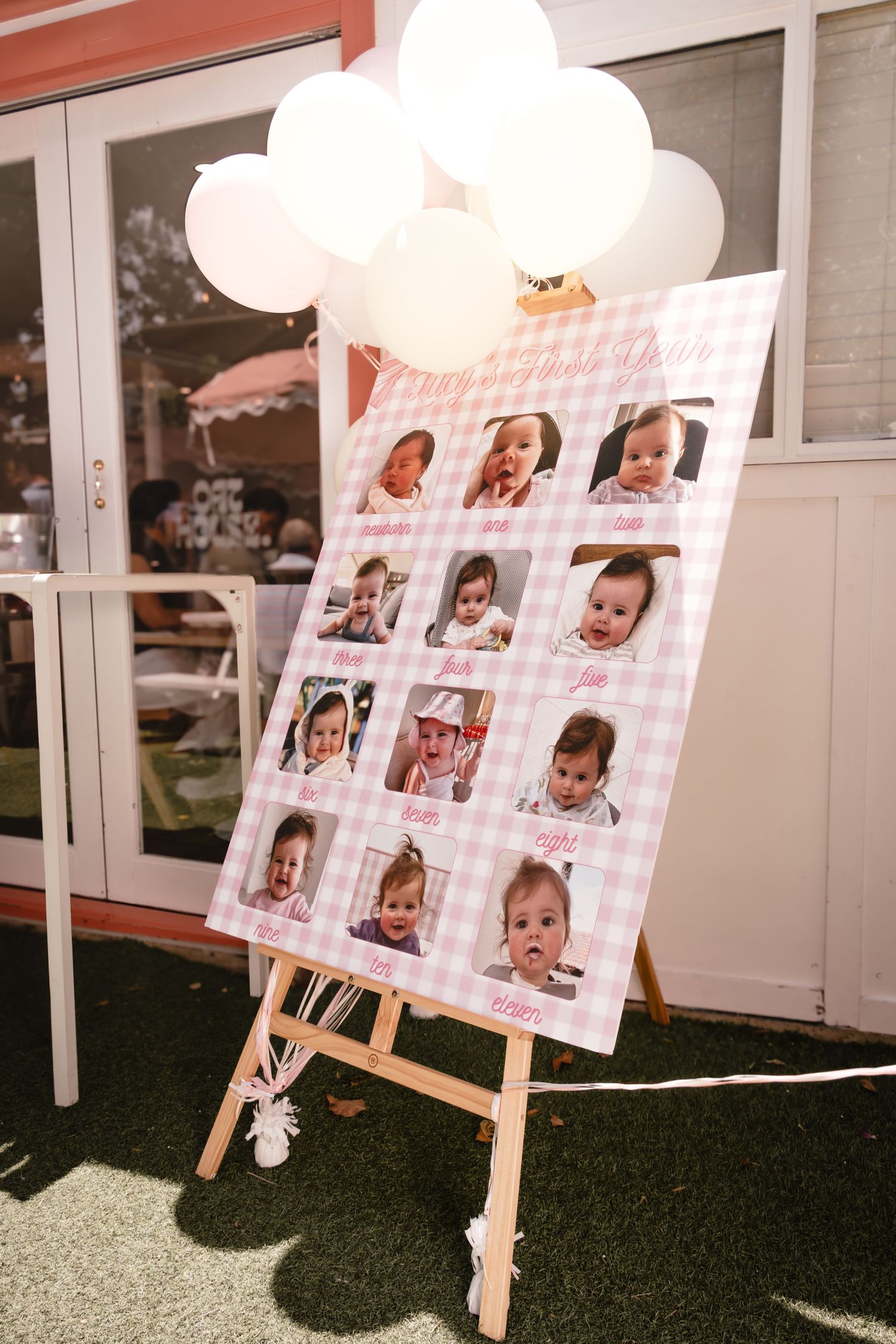 Pink gingham baby milestone photo board on easel with white balloons celebrating a first birthday party.