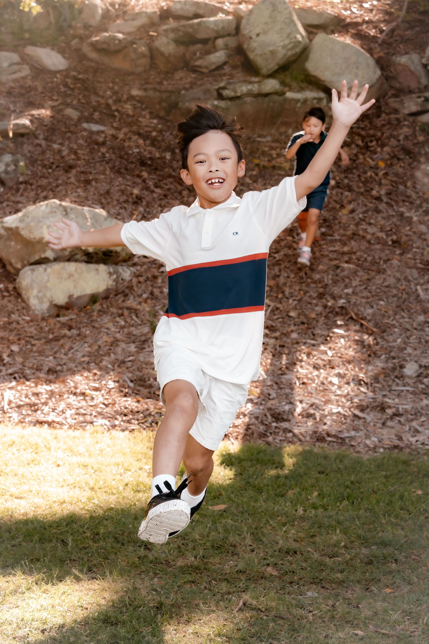 A joyful young boy in a white polo shirt runs outdoors near rocks, arms spread wide, smiling at the camera.