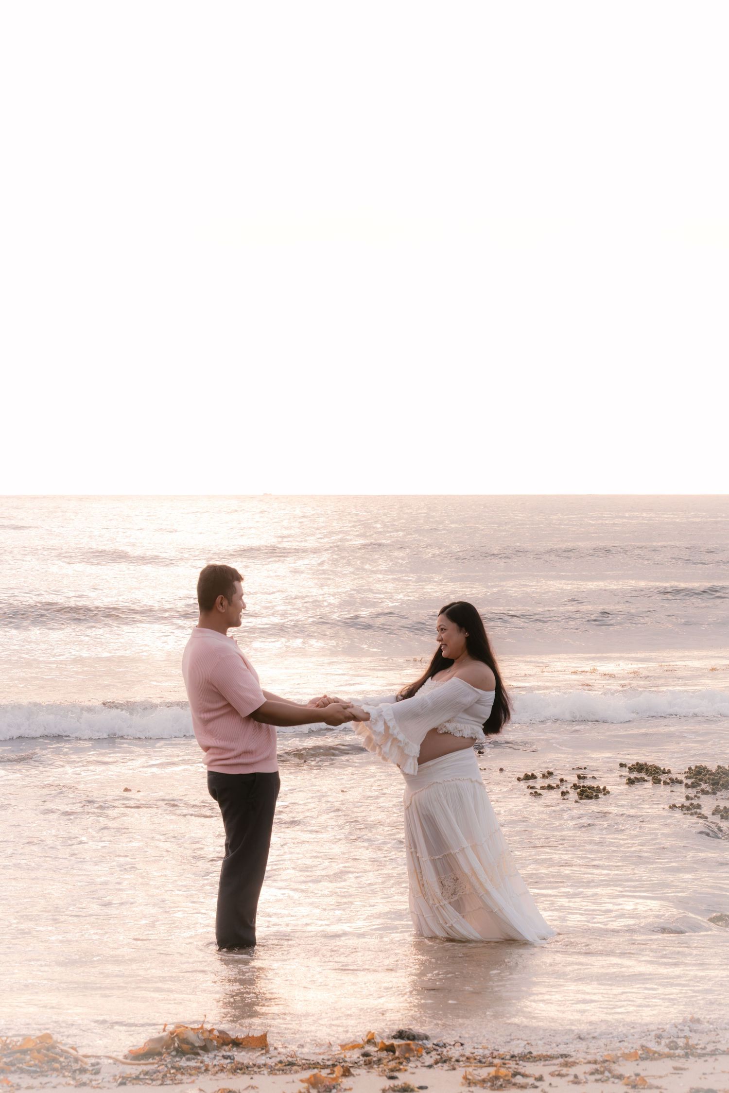 Couple holding hands on a beach at sunset, woman in white maternity dress and man in pink shirt.