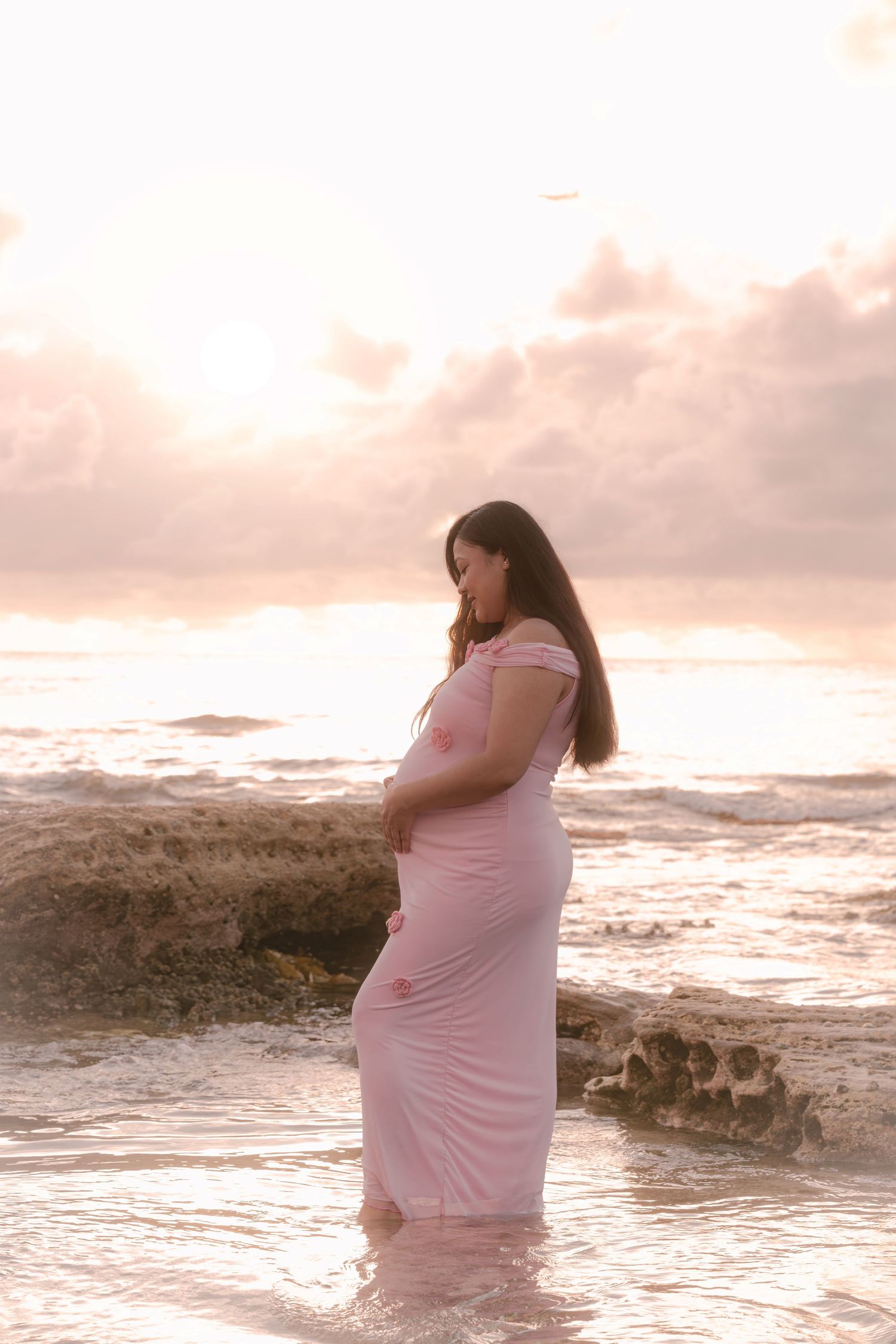 Pregnant woman in pink dress standing in ocean waves at sunset beach, cradling baby bump.