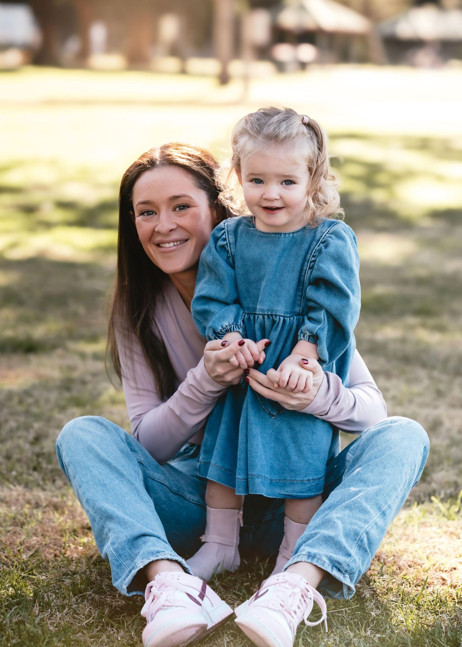A parent and toddler sitting together on the grass on a sunny day, both smiling and wearing casual clothes.
