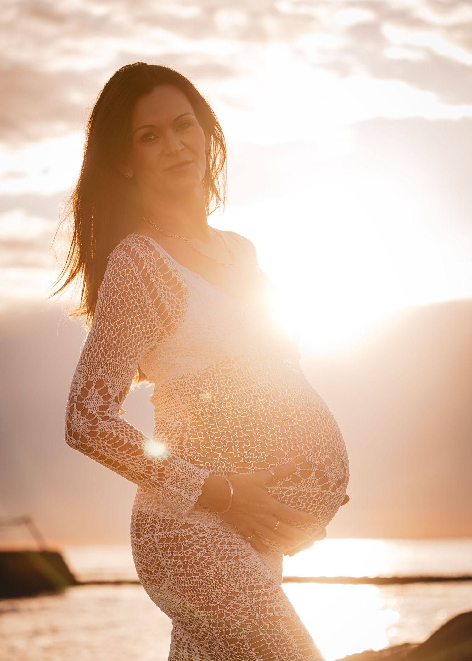 A silhouetted figure in a lace dress poses during sunset at a beach, creating a dreamy maternity photo.