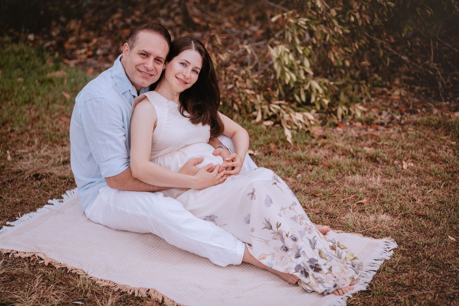 A couple in white outfits shares affectionate moments while sitting together on autumn ground during a photoshoot.