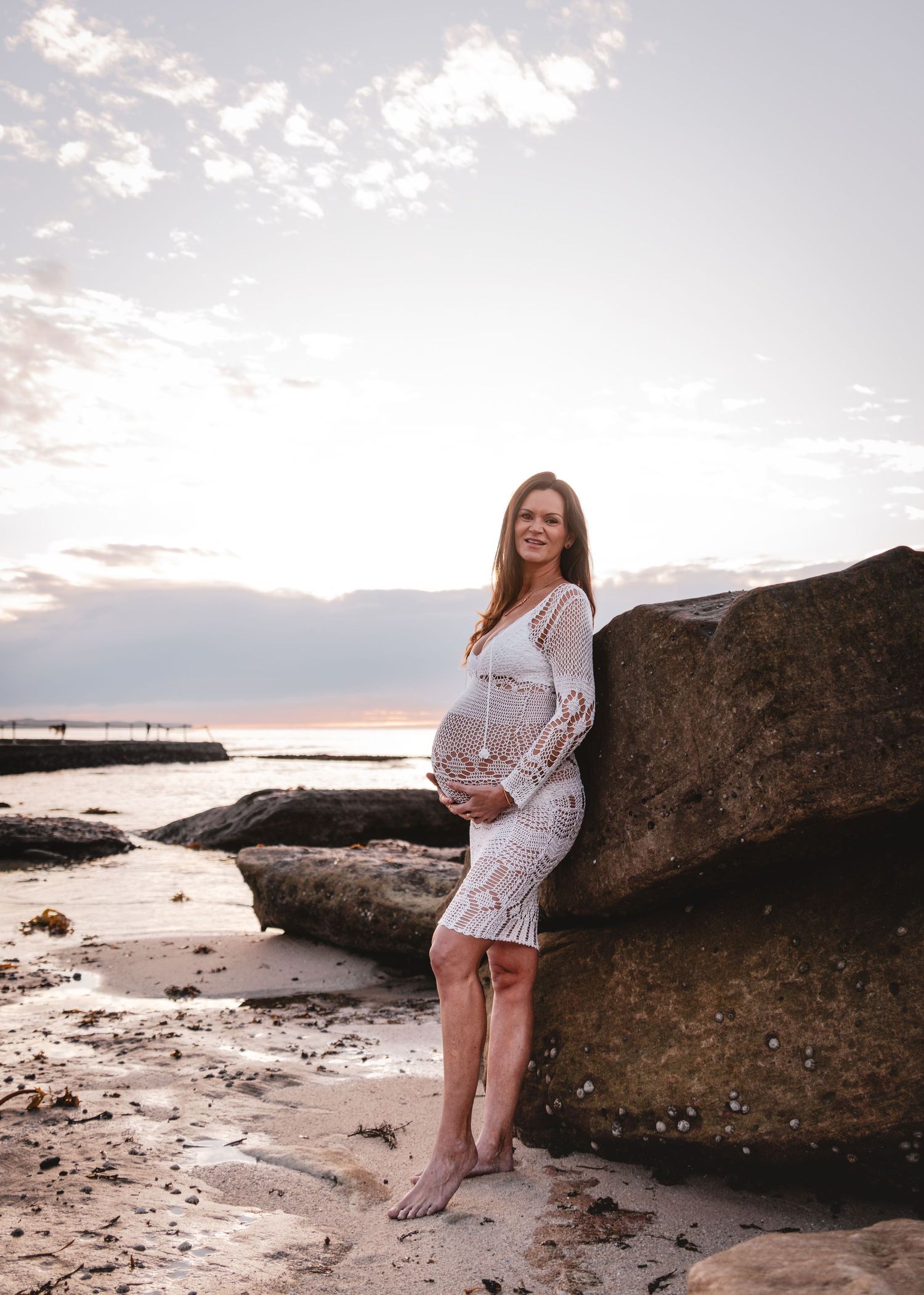 Woman in white lace dress poses on rocky beach at sunset with dramatic coastal landscape in background.