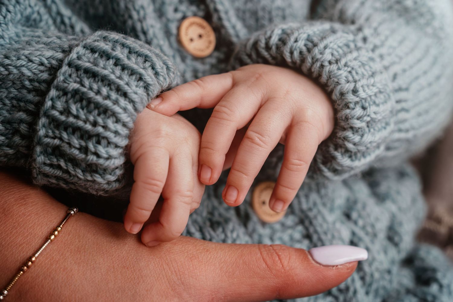 Close-up of tiny baby hands grasping grey knitted fabric against neutral background.