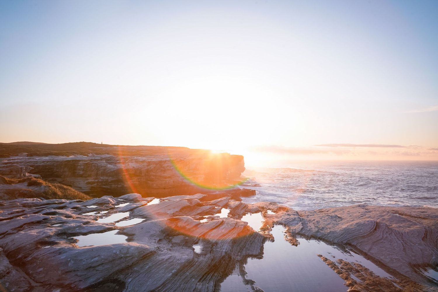 Sunrise over rocky coastal cliffs with tide pools reflecting golden light against a misty ocean horizon.