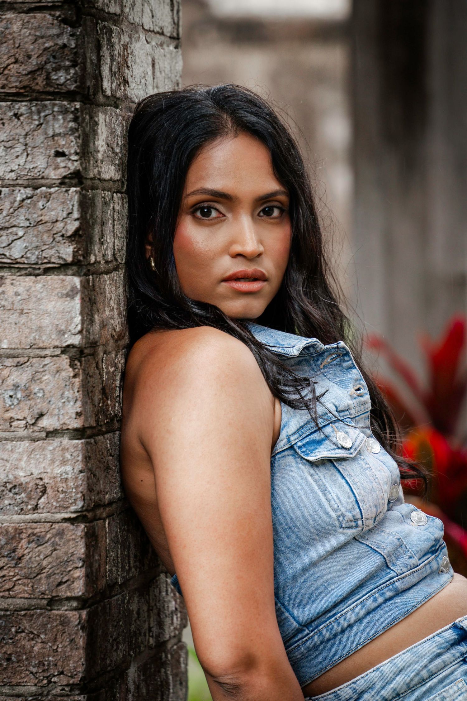 A portrait against a brick wall shows a woman in a denim crop top vest looking over her shoulder with long dark hair.