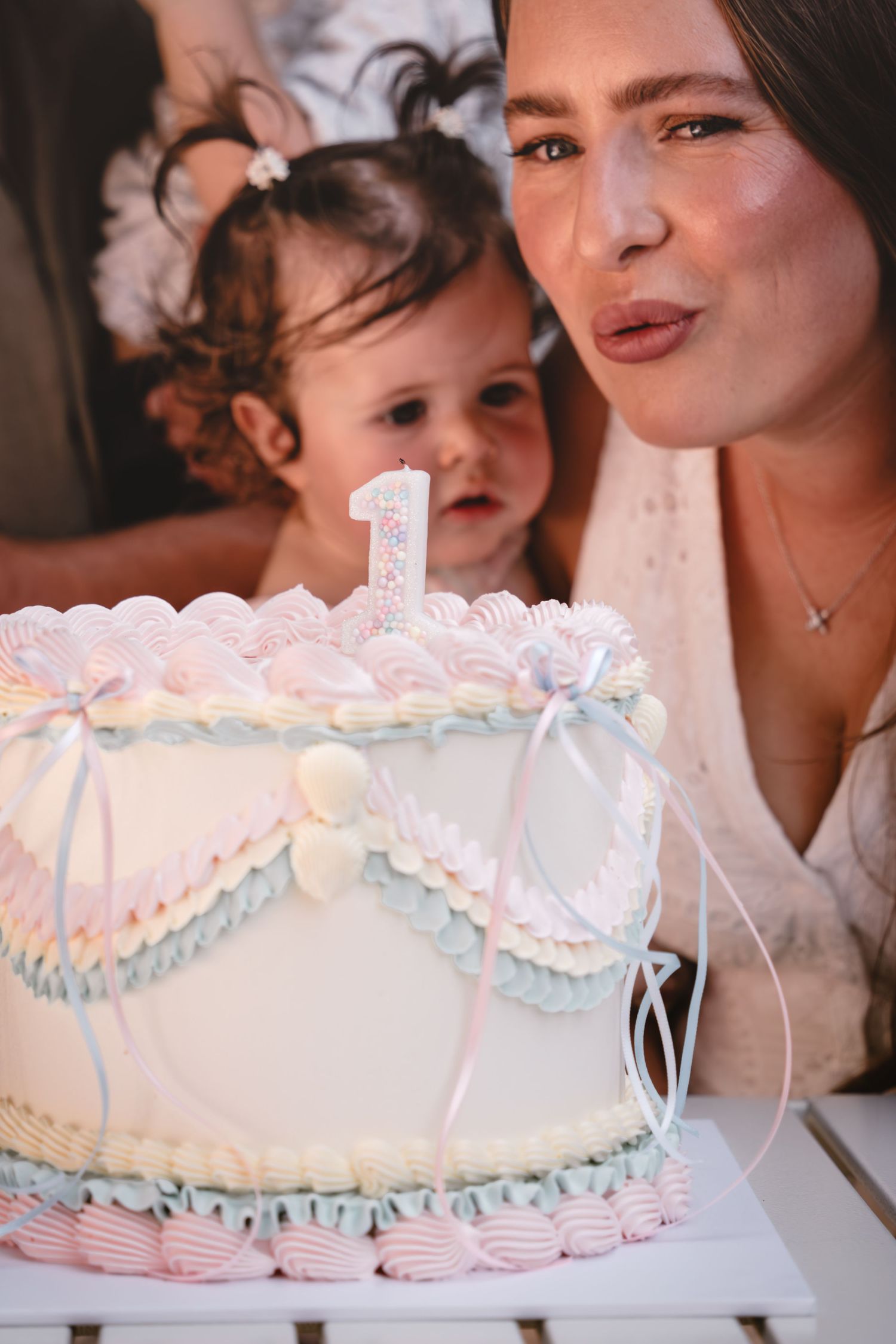 A woman blows out a candle on a decorated first birthday cake while a baby watches the celebration.