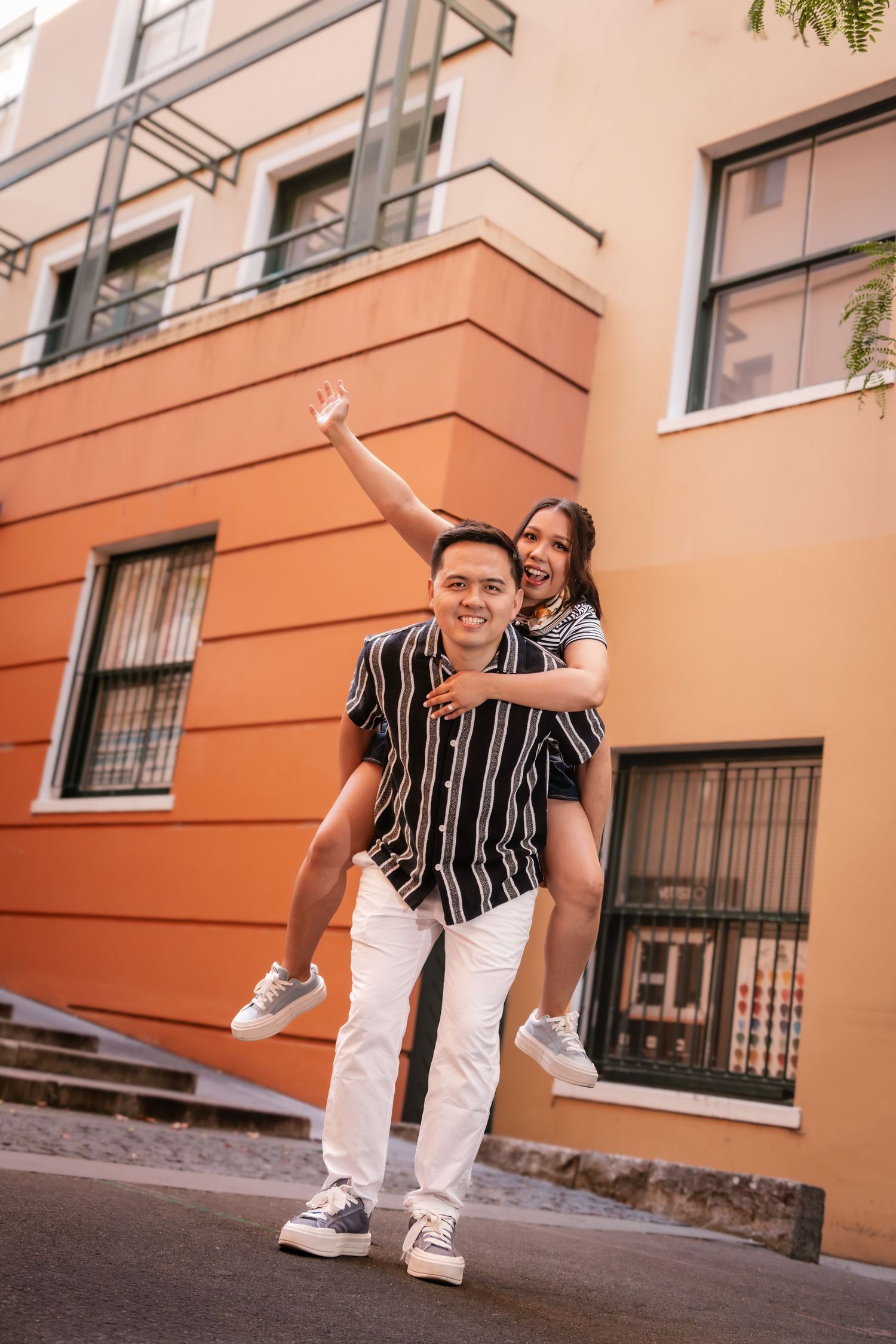 A joyful couple poses outdoors, man giving woman a piggyback ride, both smiling near a colorful orange building.