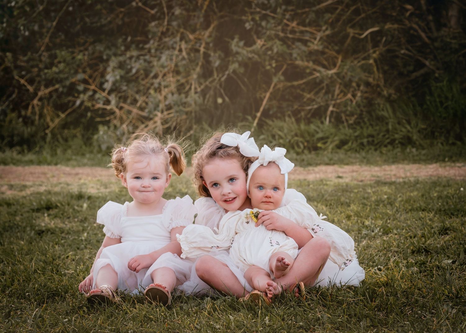 Three babies in white dresses sit together on grass with a natural backdrop.