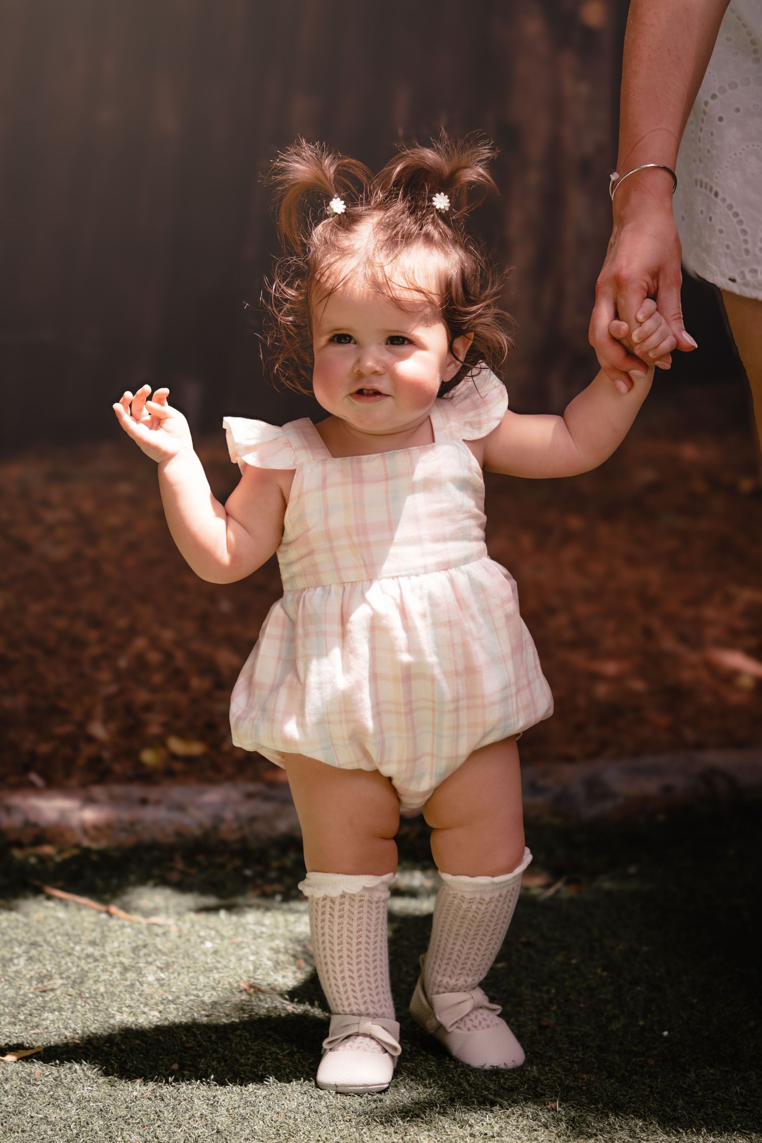 Adorable baby girl in pink plaid romper with pigtails learning to walk while holding an adult's hand outdoors.