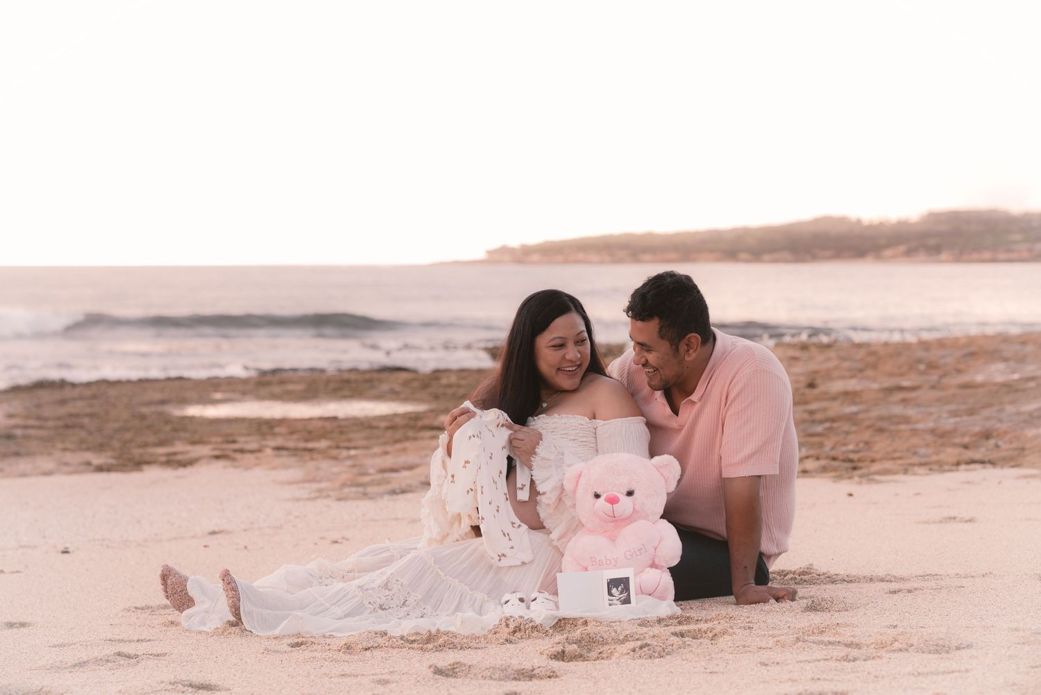 Couple sitting on beach at sunset with pink teddy bear holding ultrasound photo for maternity announcement.