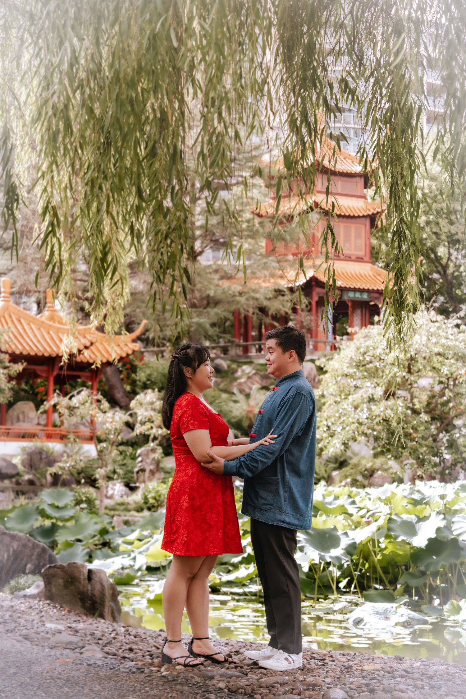 Couple in red dress and blue shirt embrace by lotus pond under willow trees at Chinese garden pagoda.