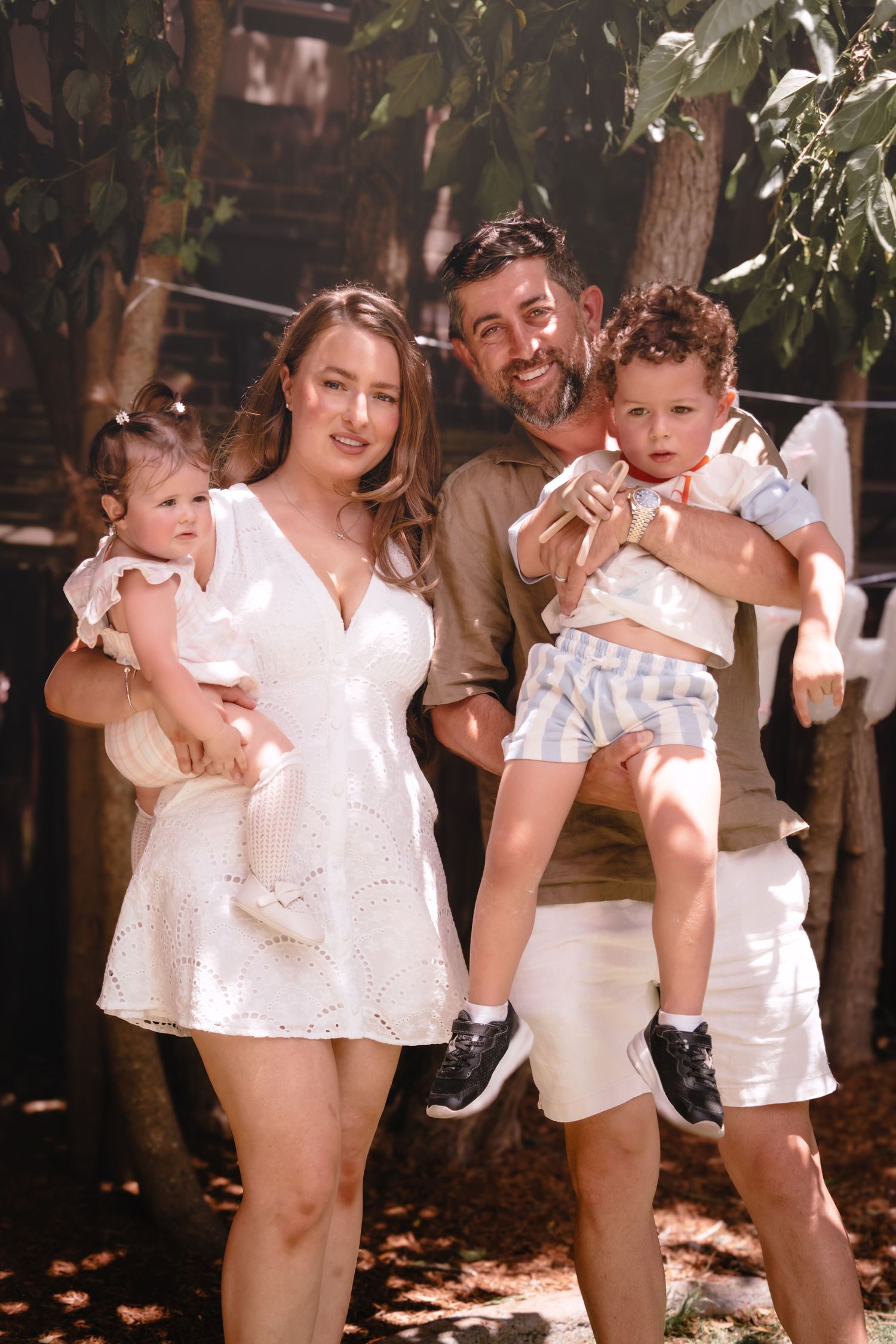 Happy family of four posing outdoors, parents holding two young children, surrounded by lush green trees in warm sunlight.