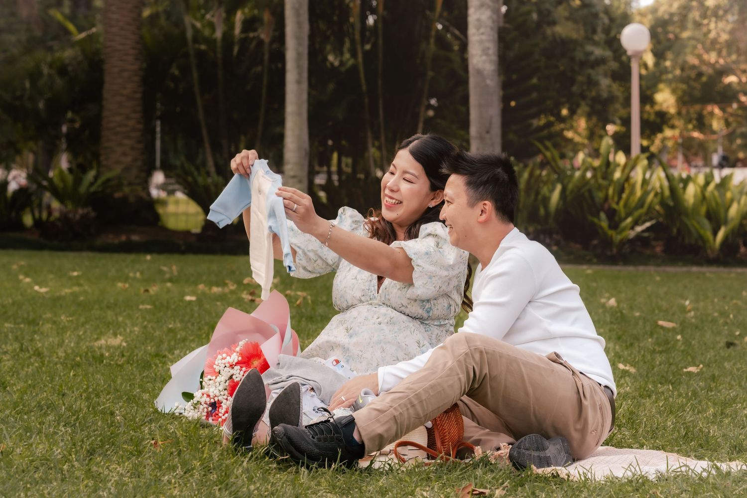 Couple sitting on grass in a park, smiling while holding up a baby onesie, with flowers nearby.